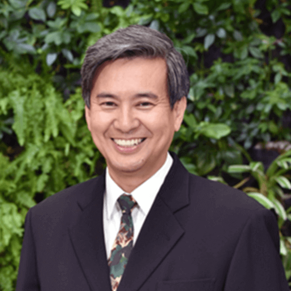 a person wearing a black suit and a tie with a camouflage pattern, standing in front of a green, leafy background