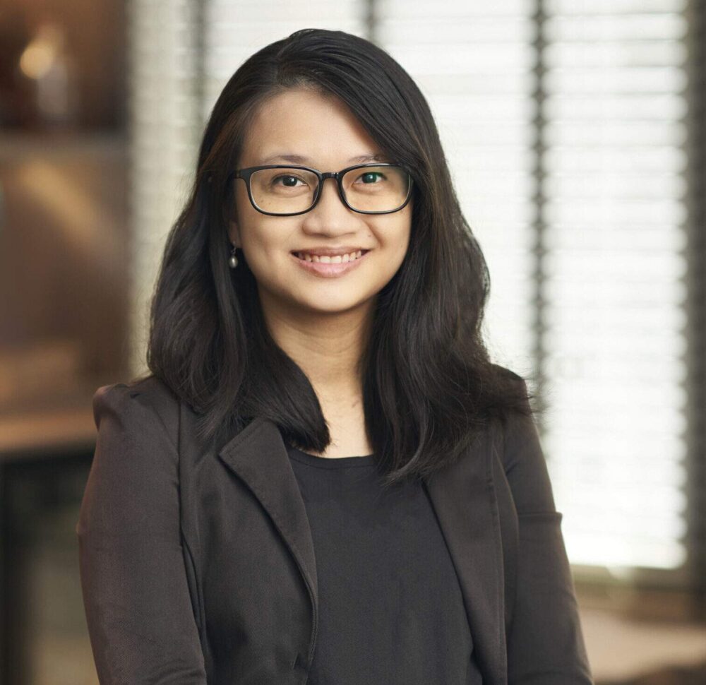 a woman with long dark hair wearing a black blazer over a black top, standing in an office setting with venetian blinds in the background