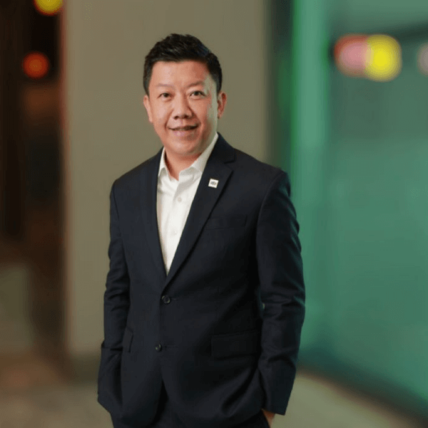 a person in a dark-colored suit with a white shirt standing in an indoor space with soft lighting in the background