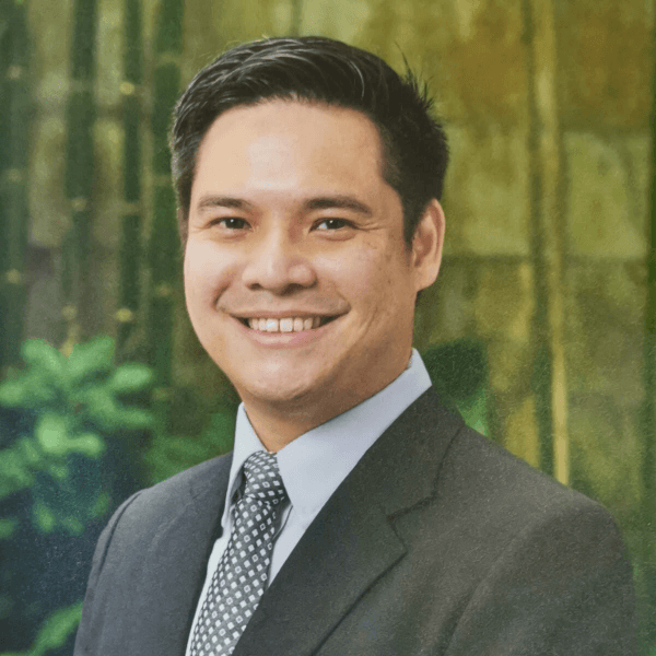 a person dressed in a business suit with a polka dot tie standing in front of a blurred green background suggestive of foliage