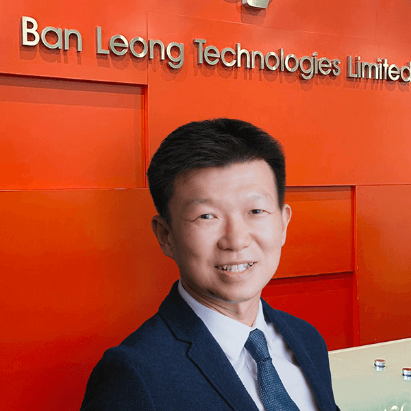 a person in a dark suit standing in front of an orange wall with "Ban Leong Technologies Limited" signage